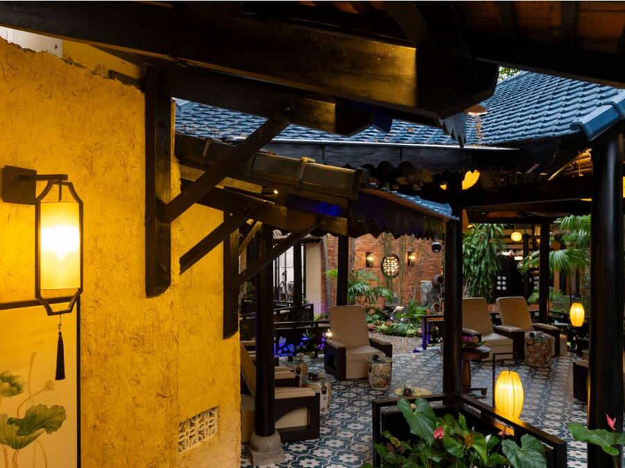 Un espacio al aire libre acogedor con muebles de madera y faroles, rodeado de plantas y un muro de ladrillo.