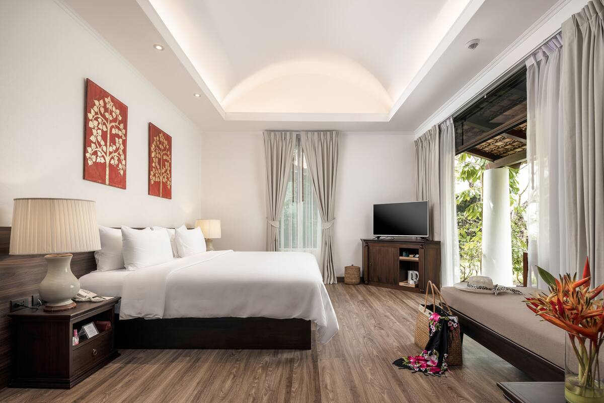 A modern hotel room with a large bed, a TV, and a view of greenery outside.