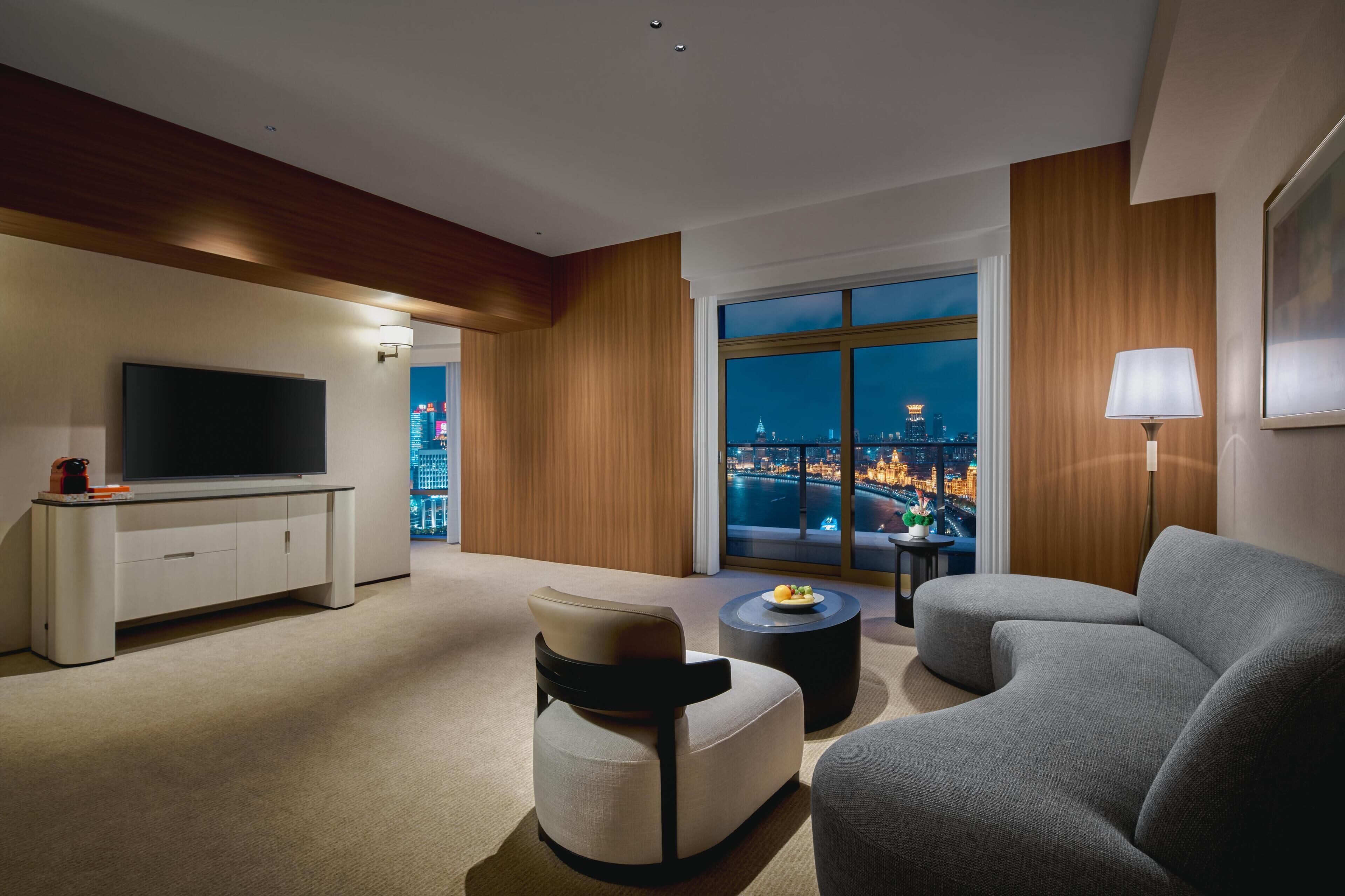 Habitación de hotel moderna con un ventanal grande que ofrece vistas a la ciudad, un televisor de pantalla plana, un sofá, una mesita de centro y una lámpara.