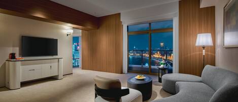 Habitación de hotel moderna con un ventanal grande que ofrece vistas a la ciudad, un televisor de pantalla plana, un sofá, una mesita de centro y una lámpara.