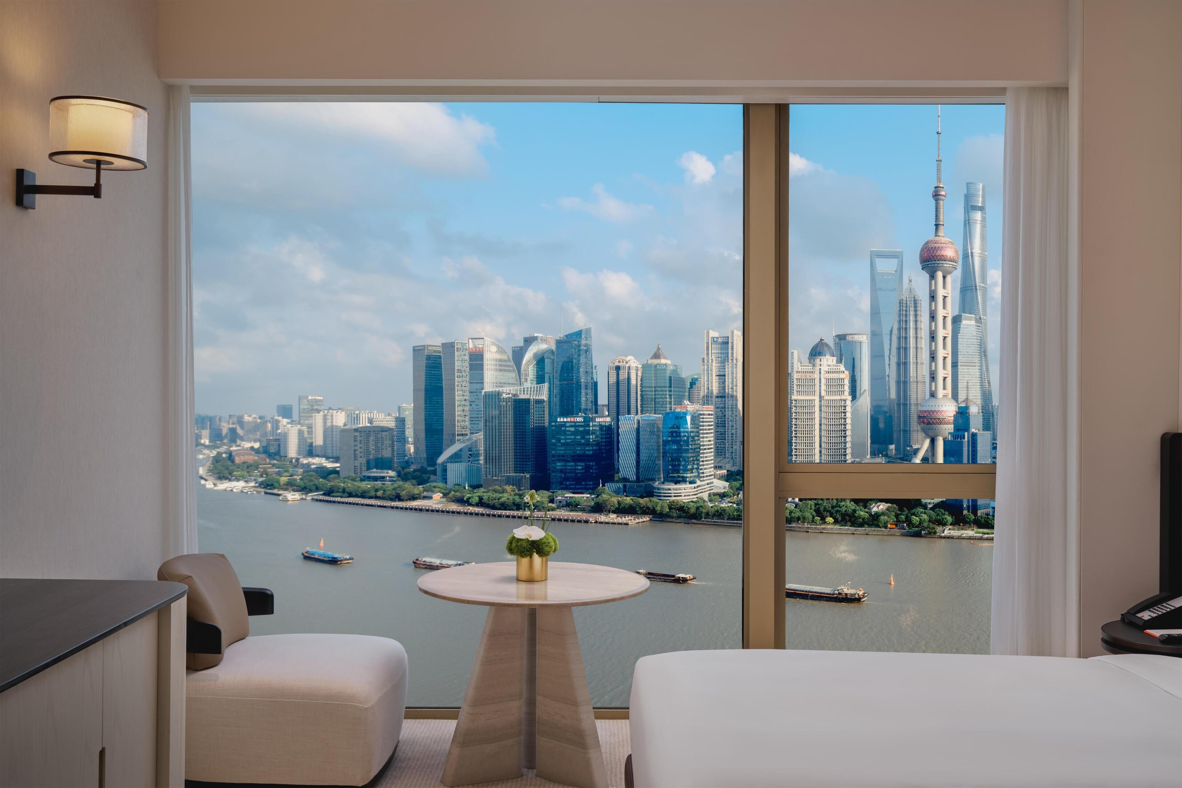A hotel room with a large window offering a view of a city skyline, a small round table with a flower vase, and a sofa.