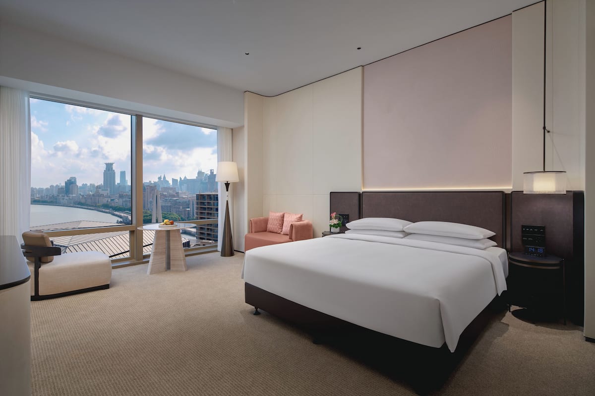 Una moderna camera d'hotel con un letto grande, una zona relax con divano e poltrona, un tavolino e vista sullo skyline della città attraverso ampie finestre.