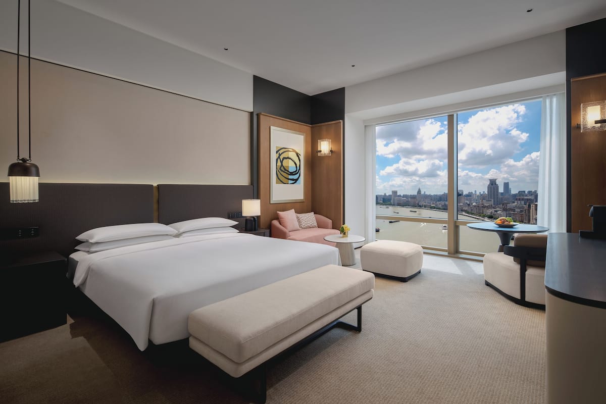A modern hotel room with a large bed, a sitting area with a sofa and armchairs, a small round table, and a view of the city through large windows.