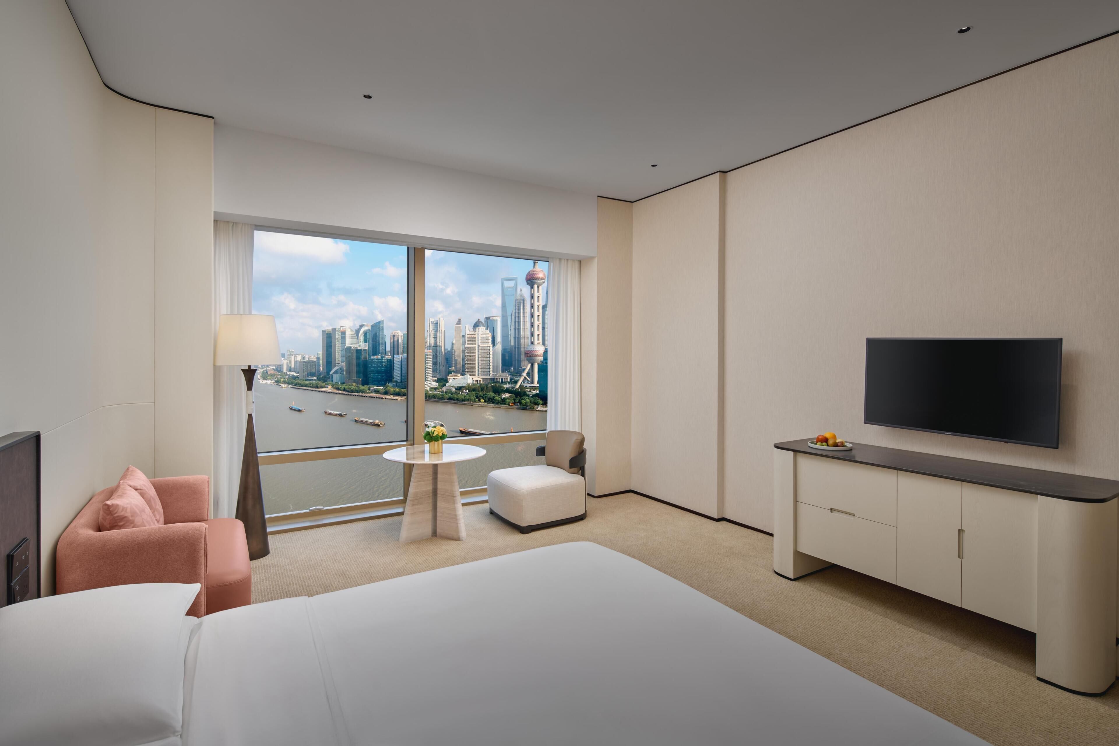 A hotel room with a large bed, a TV, a desk, a chair, and a view of a cityscape through the window.