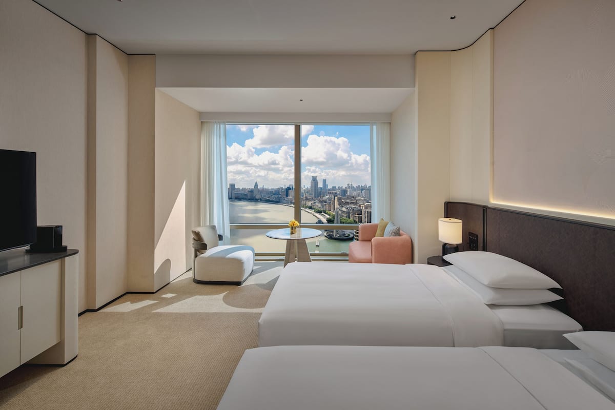 Camera d'albergo con un'ampia finestra vista città, un letto, una scrivania con televisore e un tavolino con lampada.