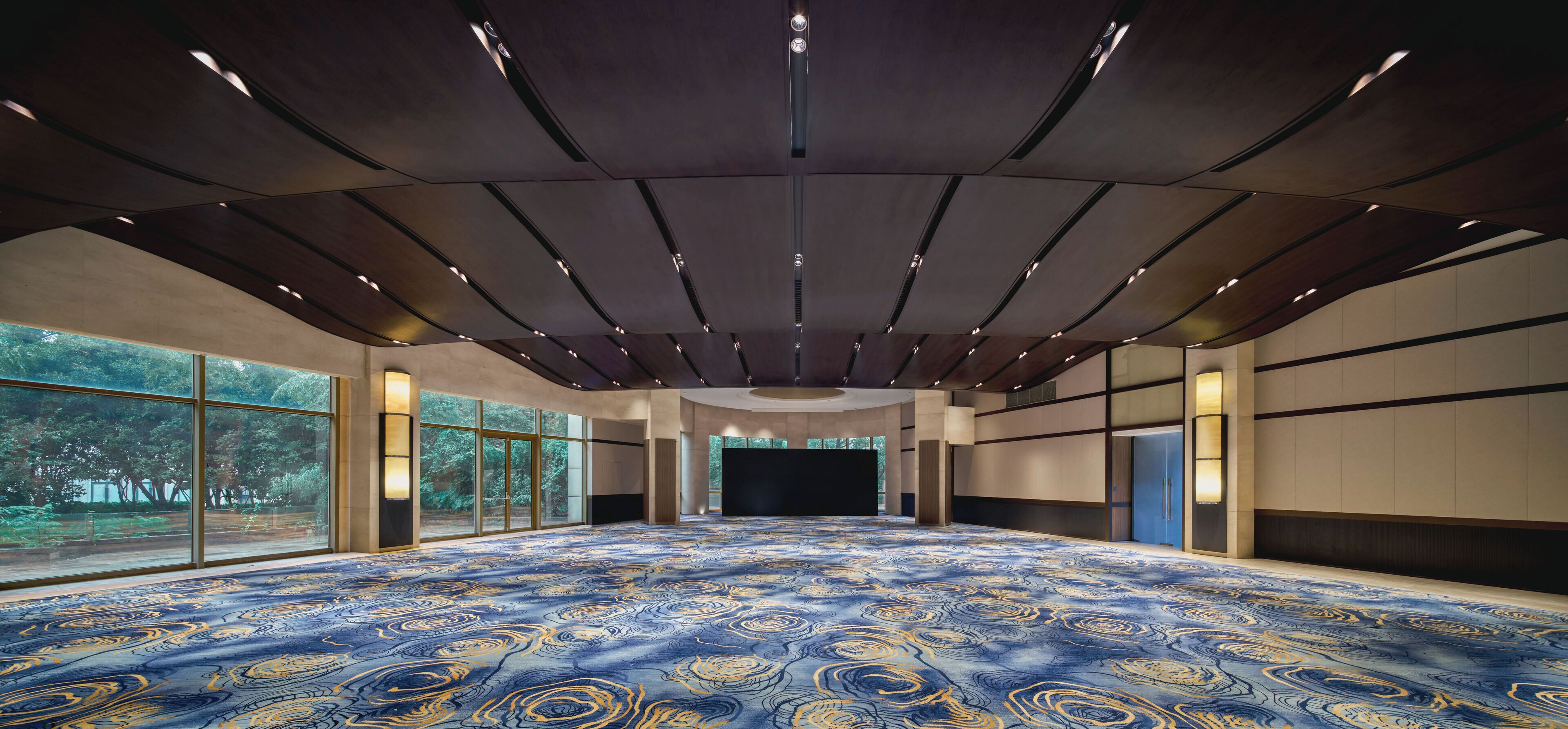 A spacious room with a patterned carpet, large windows, and a dark ceiling with recessed lighting.