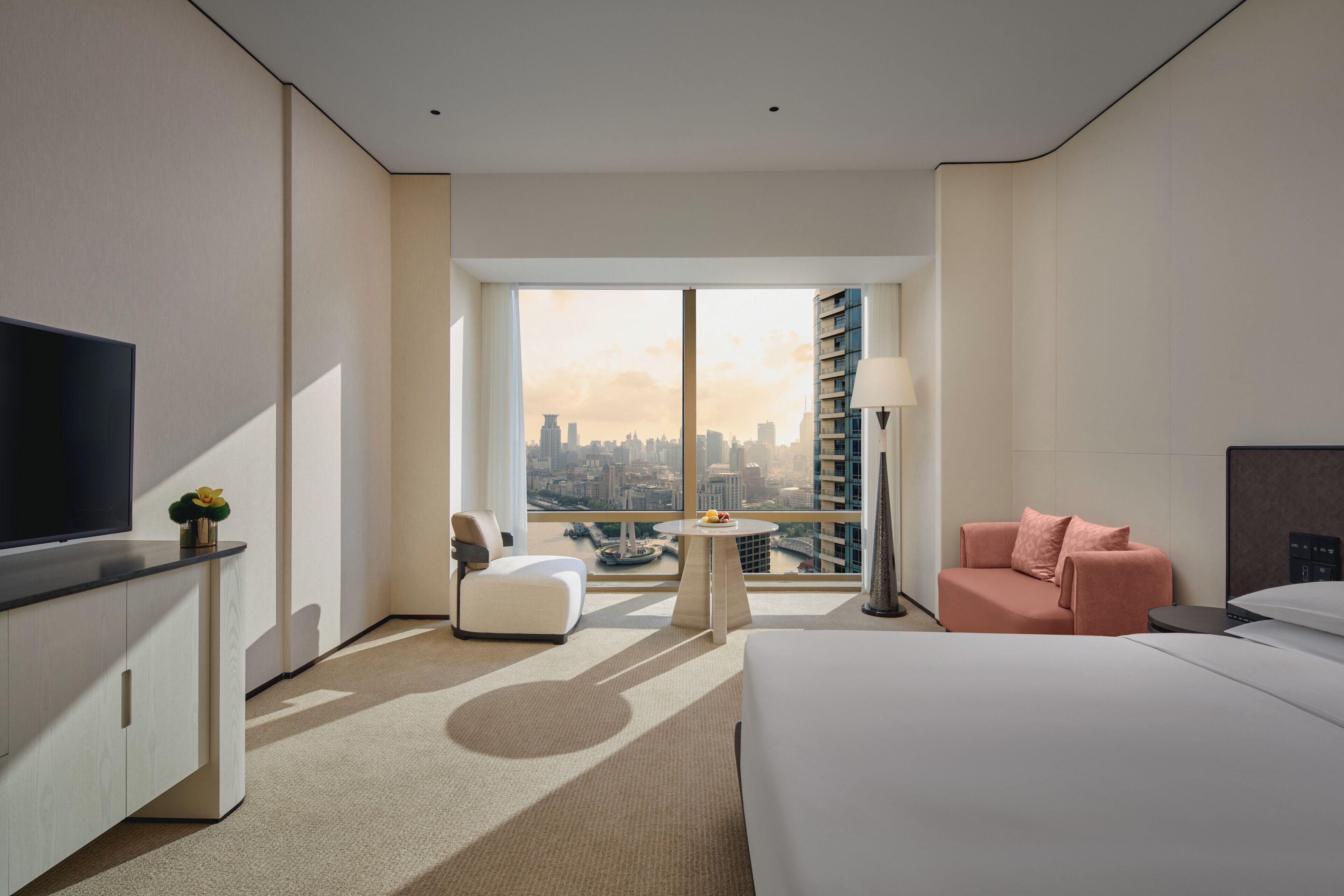 A modern hotel room with a large window, a bed, a desk, a chair, and a sofa.