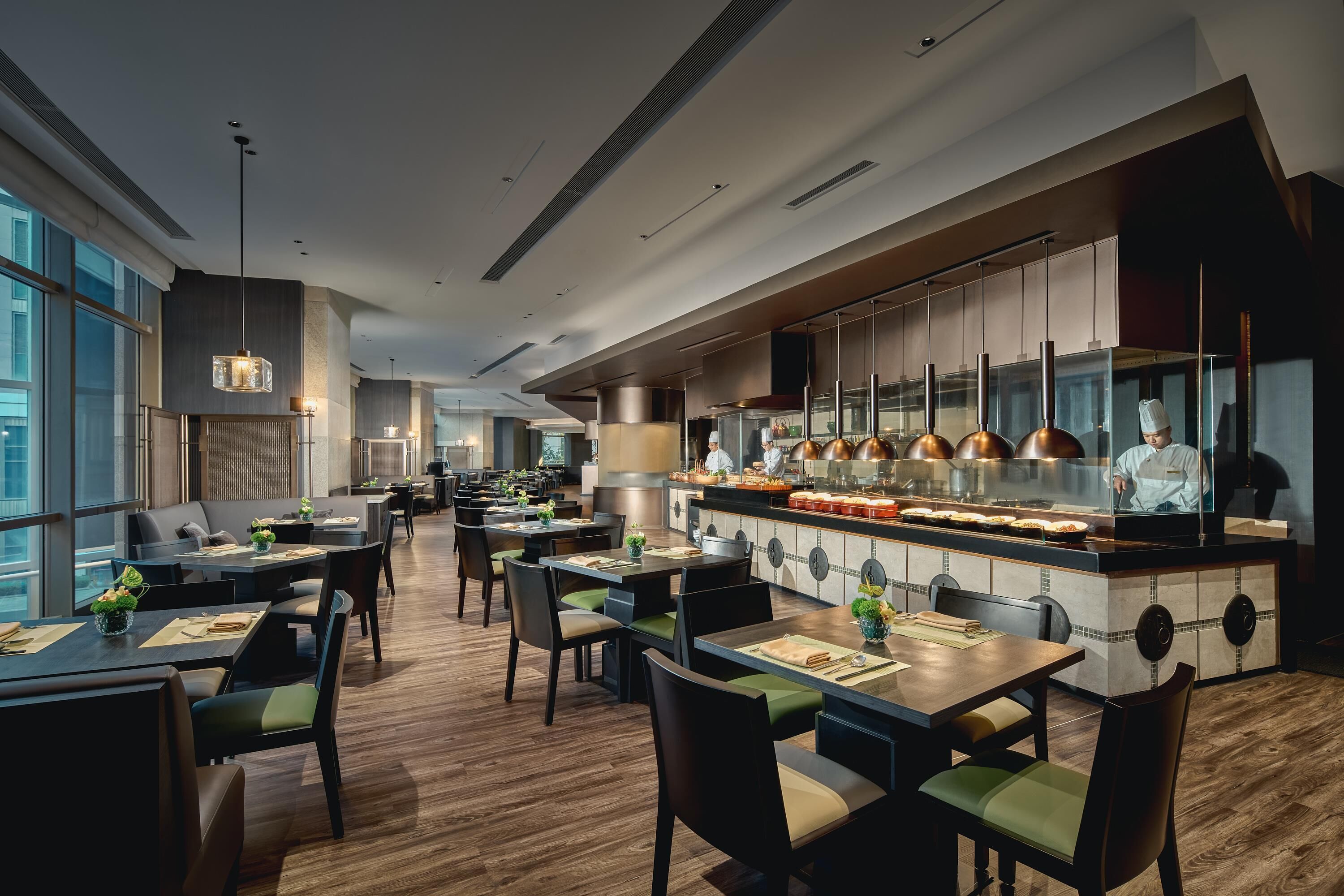 A modern restaurant with wooden flooring, green cushioned chairs, and a central counter with a chef.