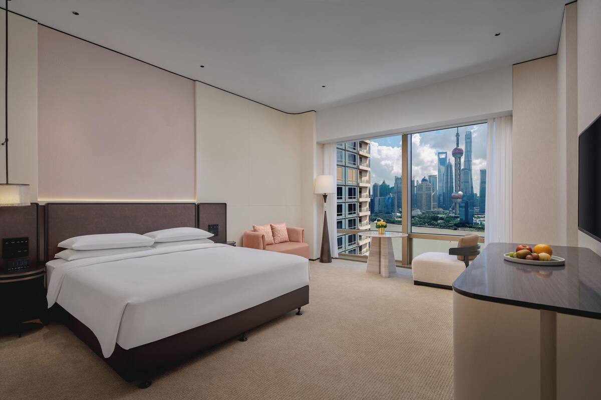 A modern hotel room with a large bed, a sofa, a desk with a fruit bowl, and a city view through large windows.