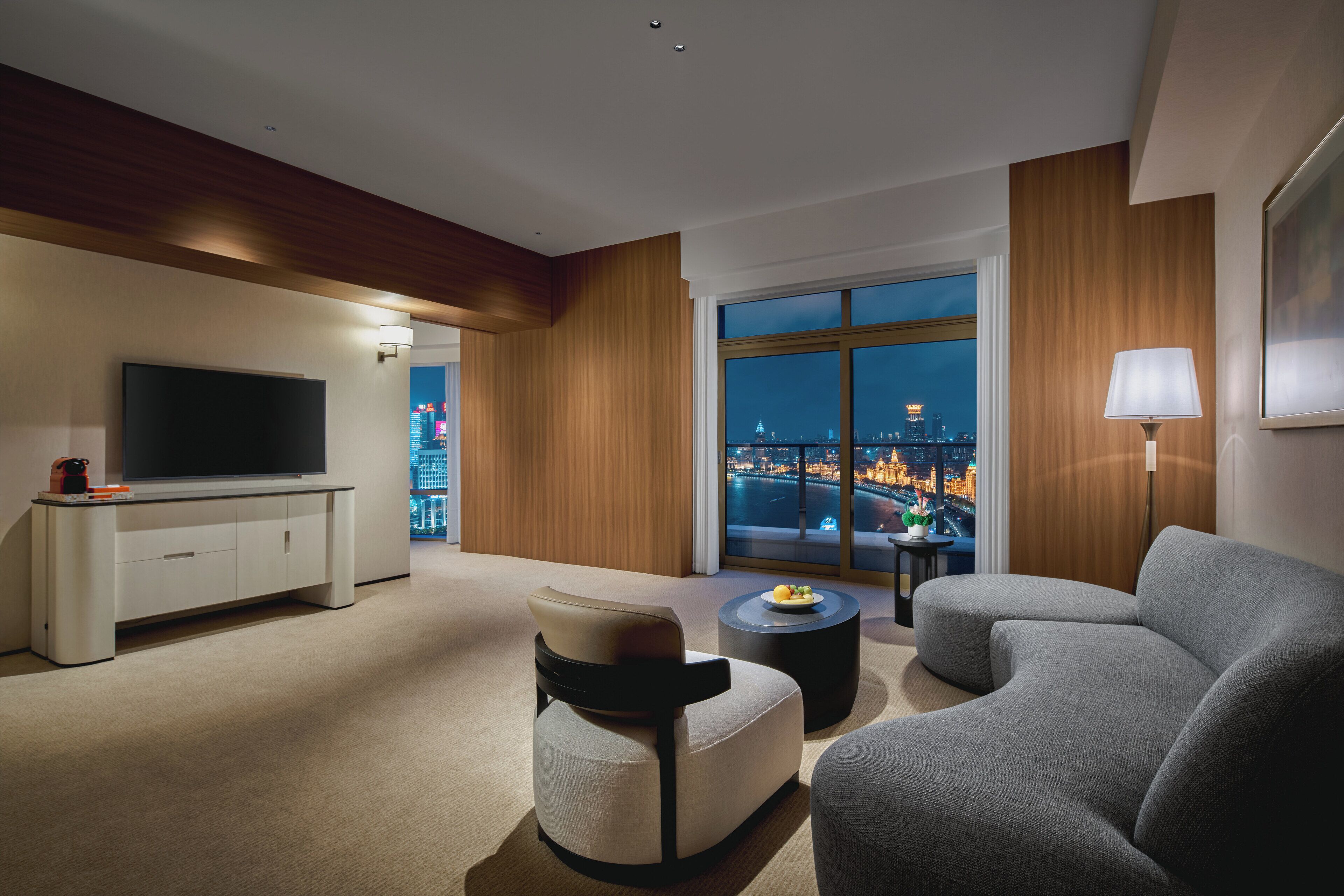 Ein modernes Hotelzimmer mit großem Fenster und Blick auf die Stadt, einem Flachbildfernseher, einem Sofa, einem kleinen Tisch und einer Lampe.