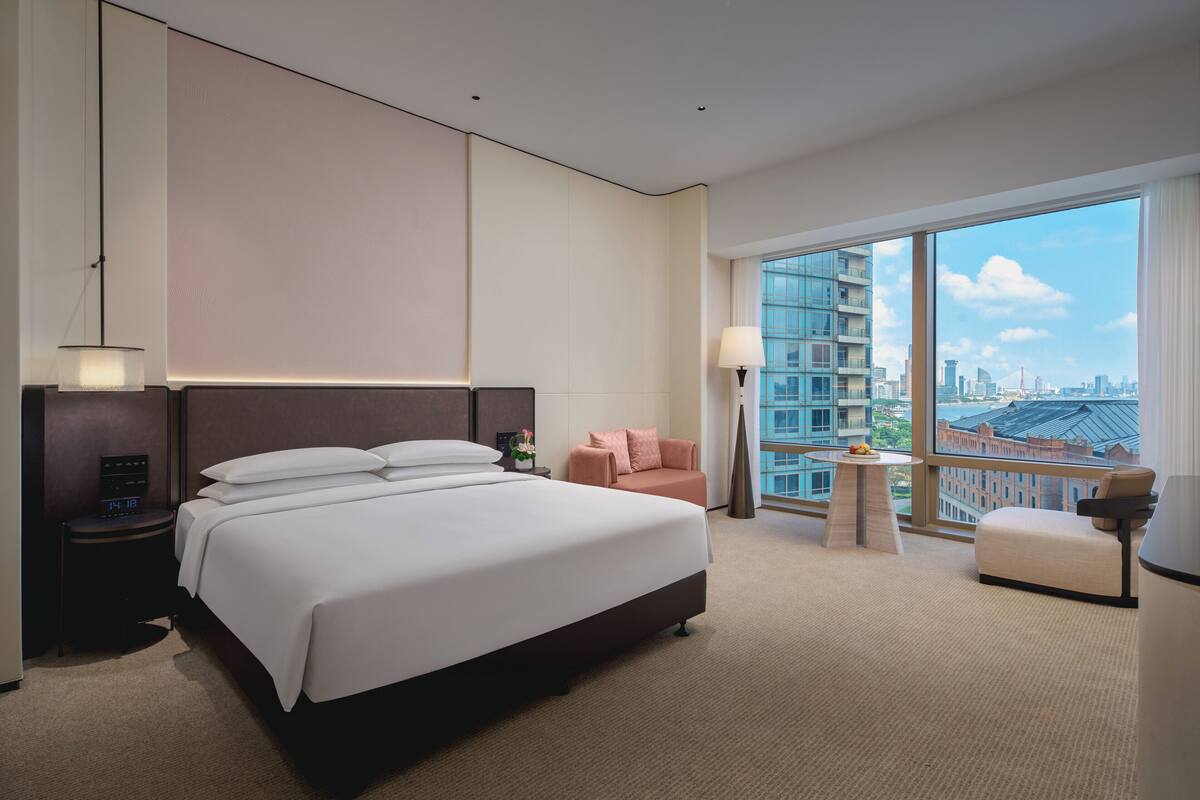 Una camera d'albergo con un letto grande, un comodino, un tavolino, una sedia e vista sulla città attraverso ampie finestre.