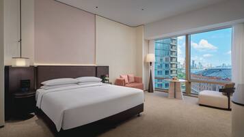 A hotel room with a large bed, a nightstand, a small table, a chair, and a view of the city through large windows.