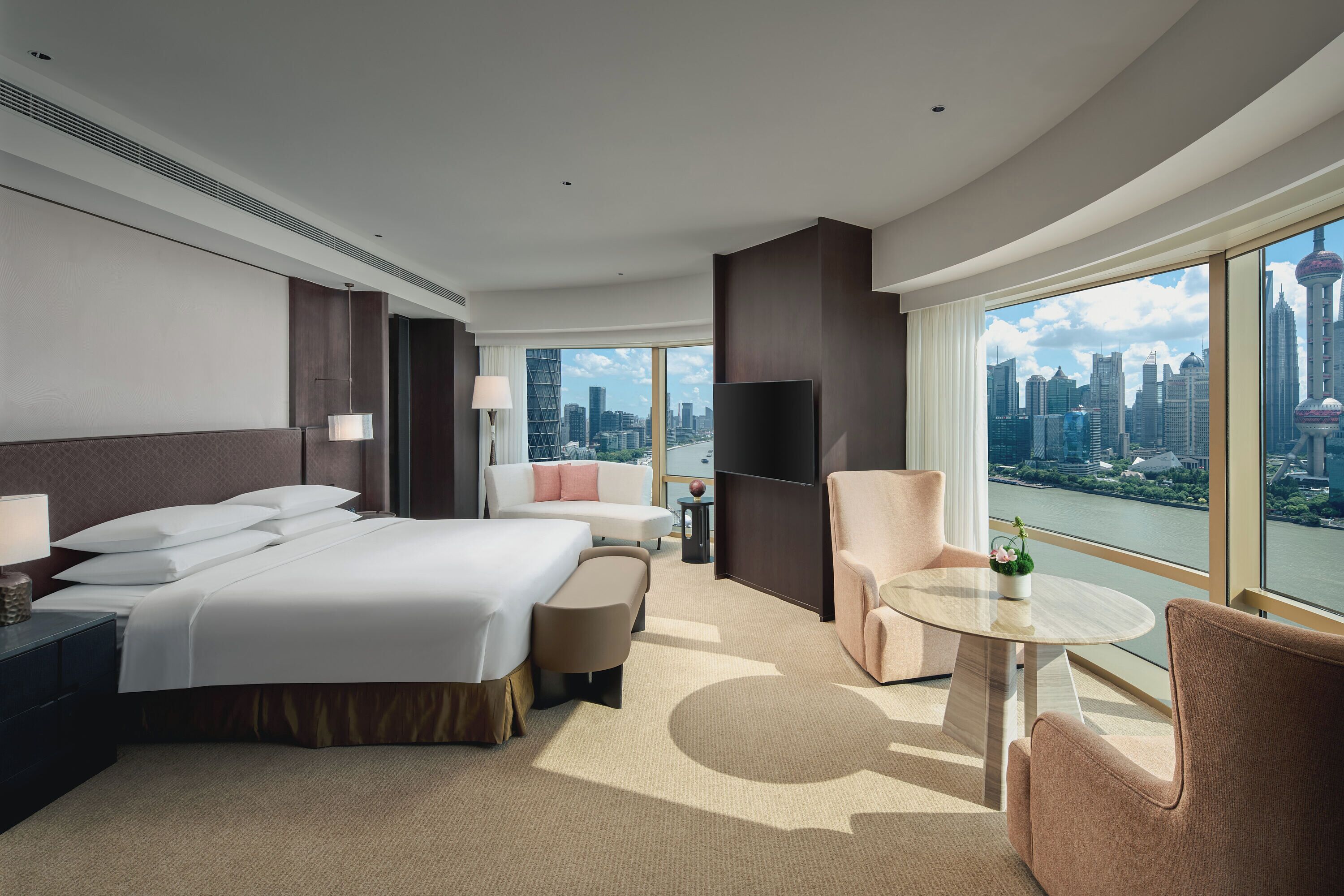 A modern hotel room with a large bed, a sofa, a small table, and a view of a cityscape through large windows.