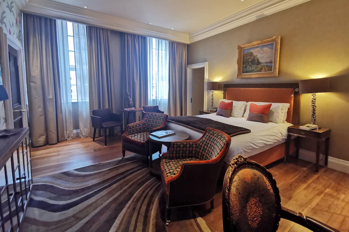 A hotel room with a large bed, a desk, two chairs, a patterned rug, and a painting on the wall.