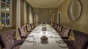 A long dining table set for a formal meal, surrounded by purple upholstered chairs, with a decorative wall piece above.