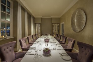 A long dining table set for a formal meal, surrounded by purple upholstered chairs, with a decorative wall piece above.