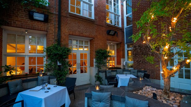 An outdoor patio with tables and chairs, a tree with lights, and a brick building in the background. - The Roseate Reading (Reading)
