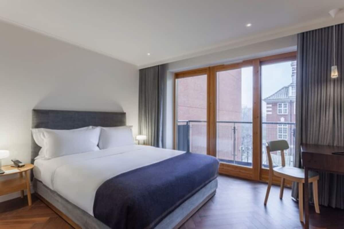 A hotel room with a large bed, a desk, and a chair. There is a window with a view of buildings outside.