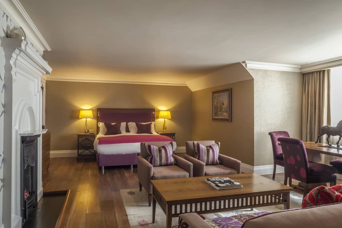 A hotel room with a bed, two armchairs, a sofa, a wooden coffee table, a fireplace, and a dining table with chairs.