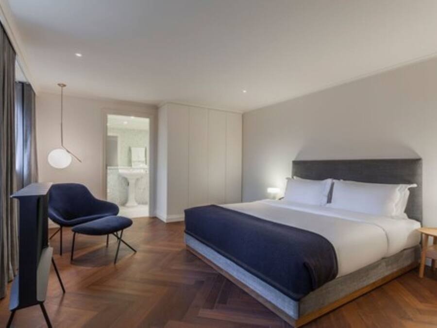 A modern hotel room with a large bed, a chair, a bedside table, and a hanging light fixture.