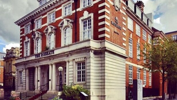 A large, red-brick building with a classical architectural style, featuring a prominent entrance with steps and columns, and multiple windows.
