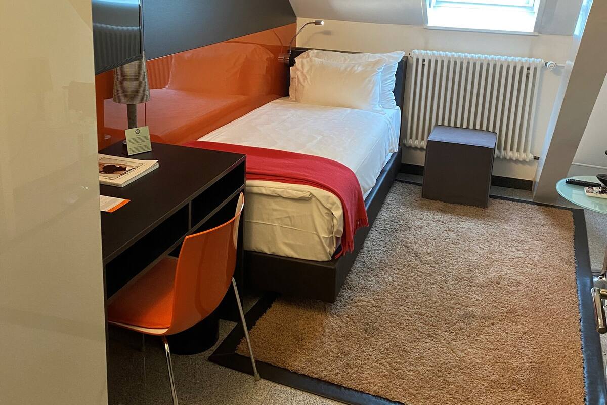 A compact hotel room with a bed, a desk, and a chair. There is a window with a view of the sky, a radiator, and a small table with a lamp.