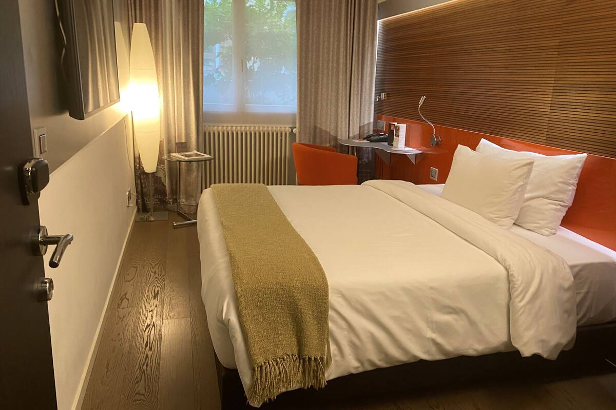 A hotel room with a large bed, a bedside table, a lamp, a window with curtains, and a wooden headboard.
