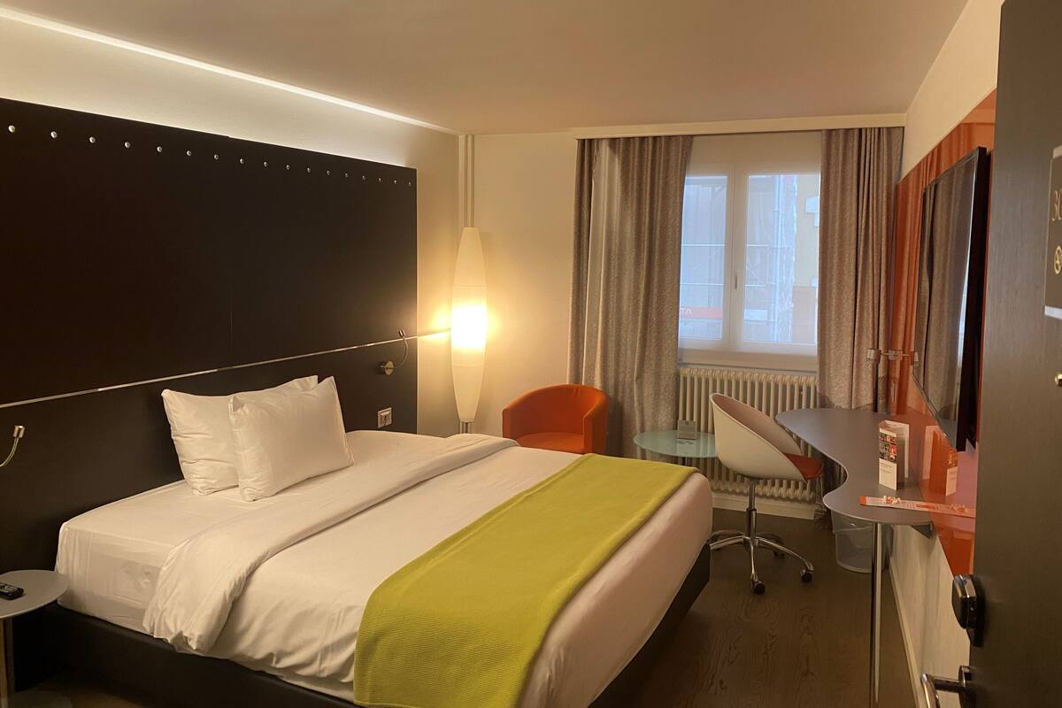 A hotel room with a bed, a desk, a chair, a lamp, and a window with curtains.