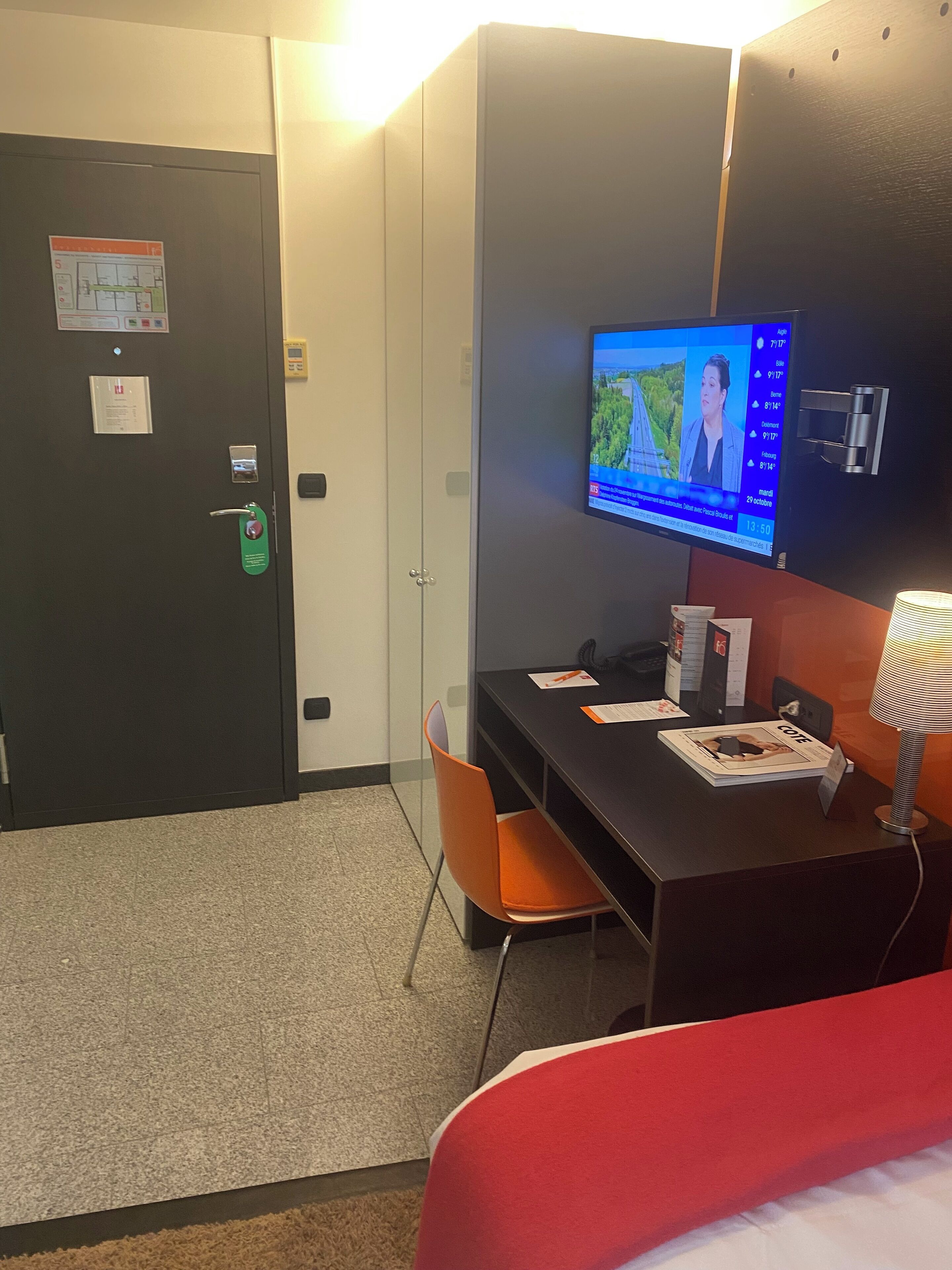 A hotel room with a desk, chair, bed, television, and a door with a security panel.