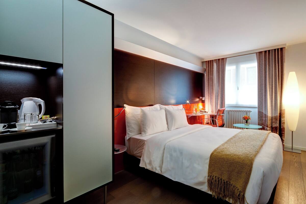 A hotel room with a bed, a desk with a coffee maker, a TV, and a window with curtains.