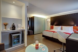 Una habitación de hotel moderna con cama, escritorio, refrigerador y una pequeña mesa con un florero y vasos.