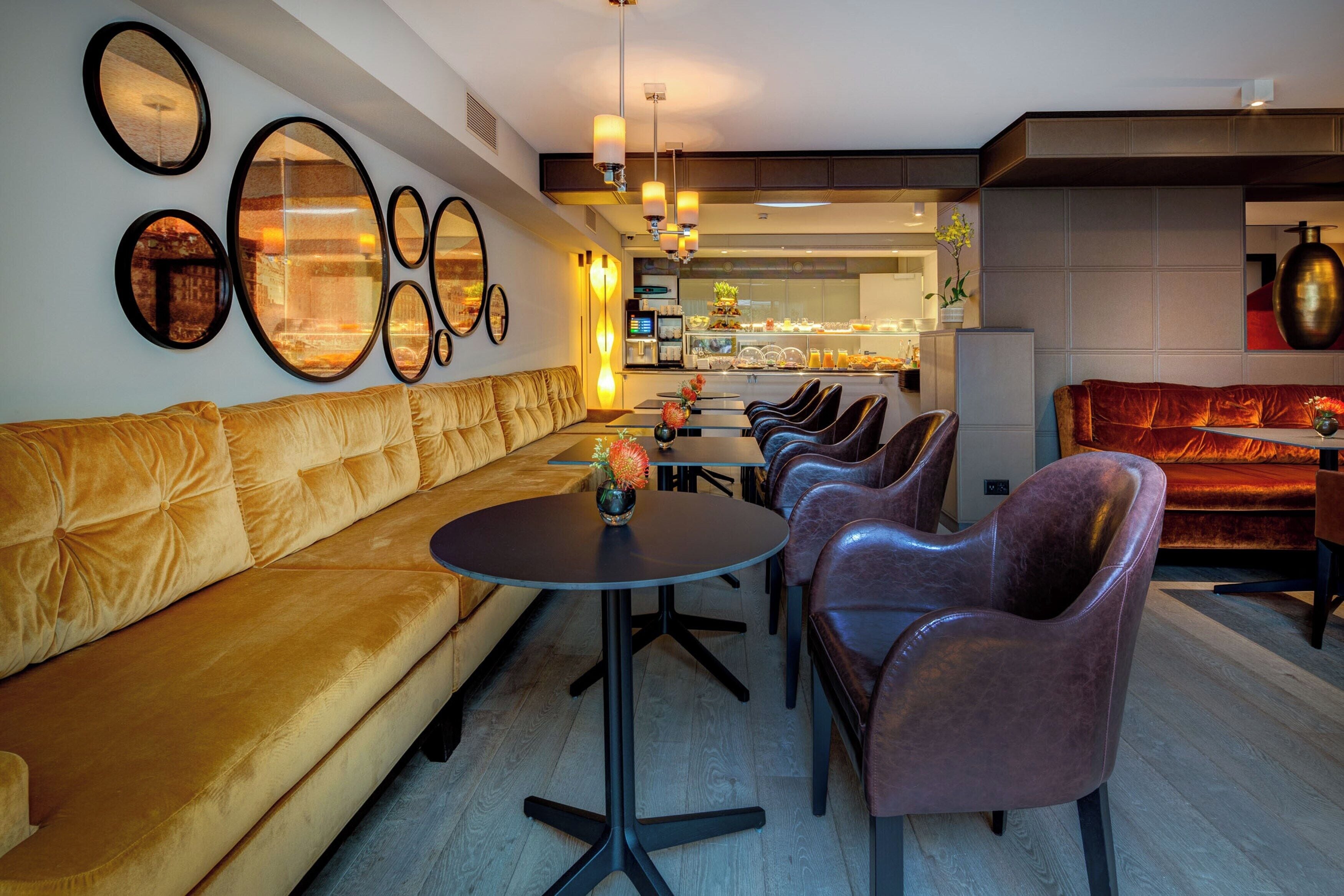 A modern lounge area with a plush gold sofa, leather chairs, and a central round table with a vase of flowers.