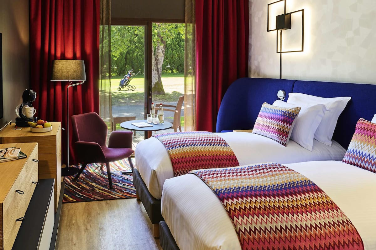A hotel room with a large bed, a desk with a coffee maker, a chair, and a view of a garden through the window.
