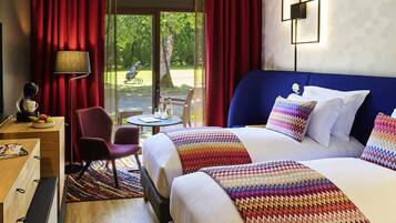 Habitación de hotel con una cama grande, un escritorio con cafetera, una silla y vista a un jardín por la ventana.