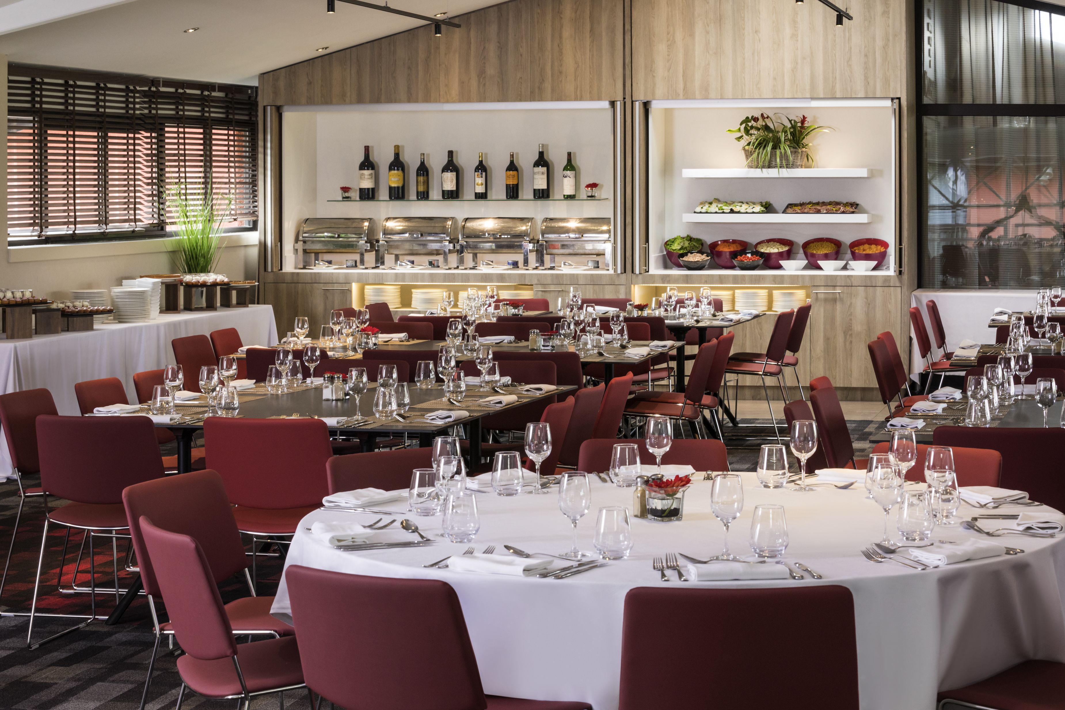 A restaurant with round tables set for dining, red chairs, and a variety of wine bottles on display.