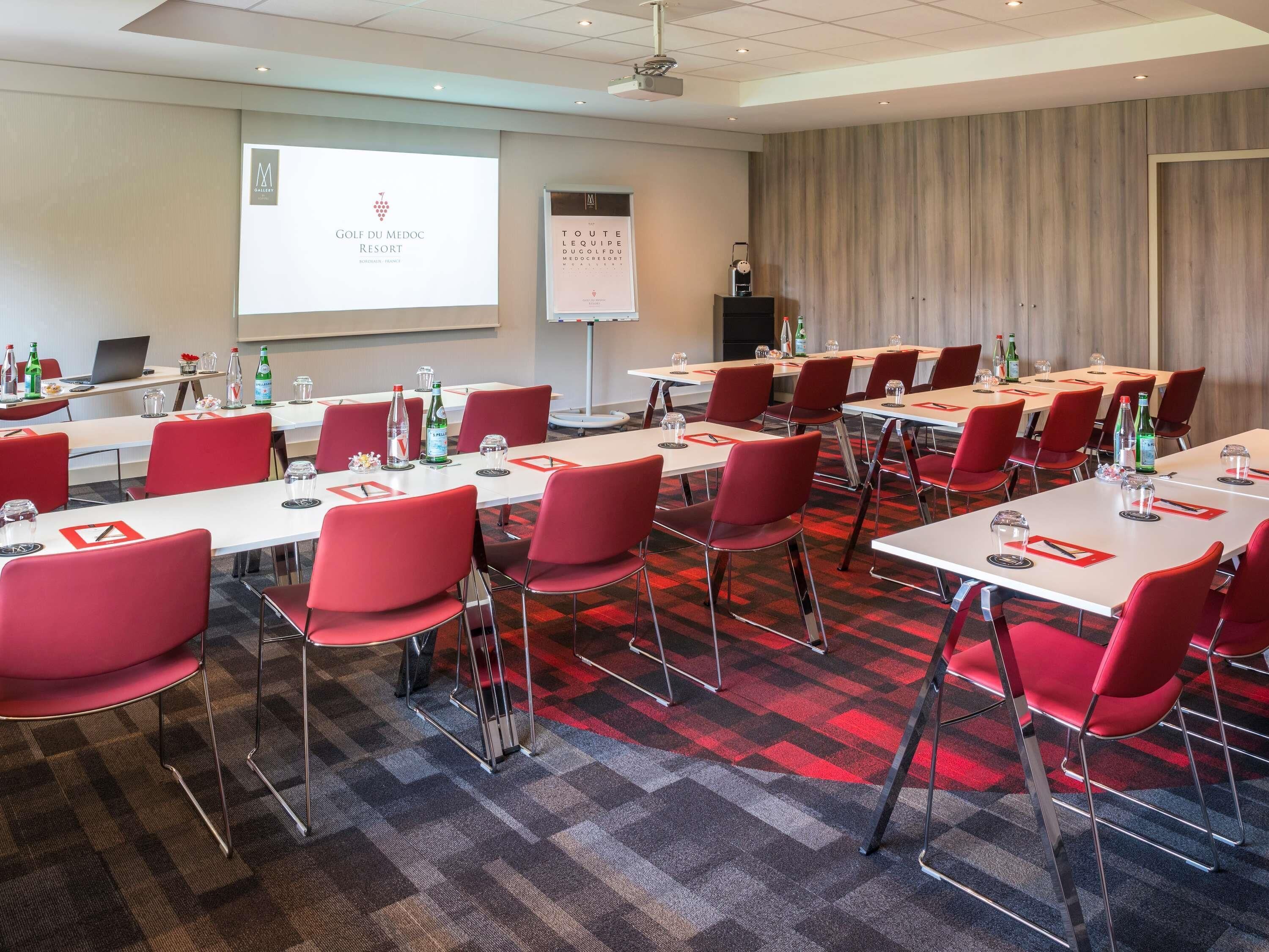 A conference room with red chairs, white tables, and a projector screen displaying a presentation.