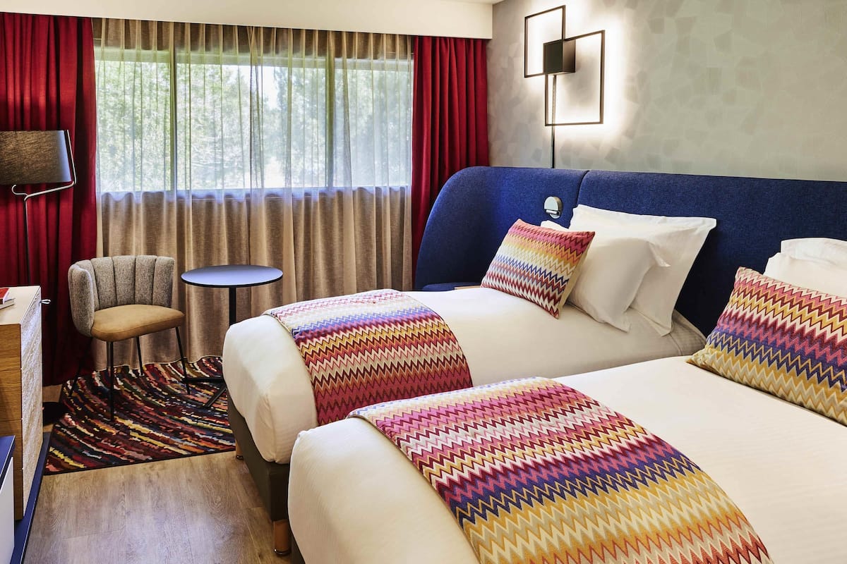 A hotel room with two beds, a chair, a small table, and a view of trees through the window.