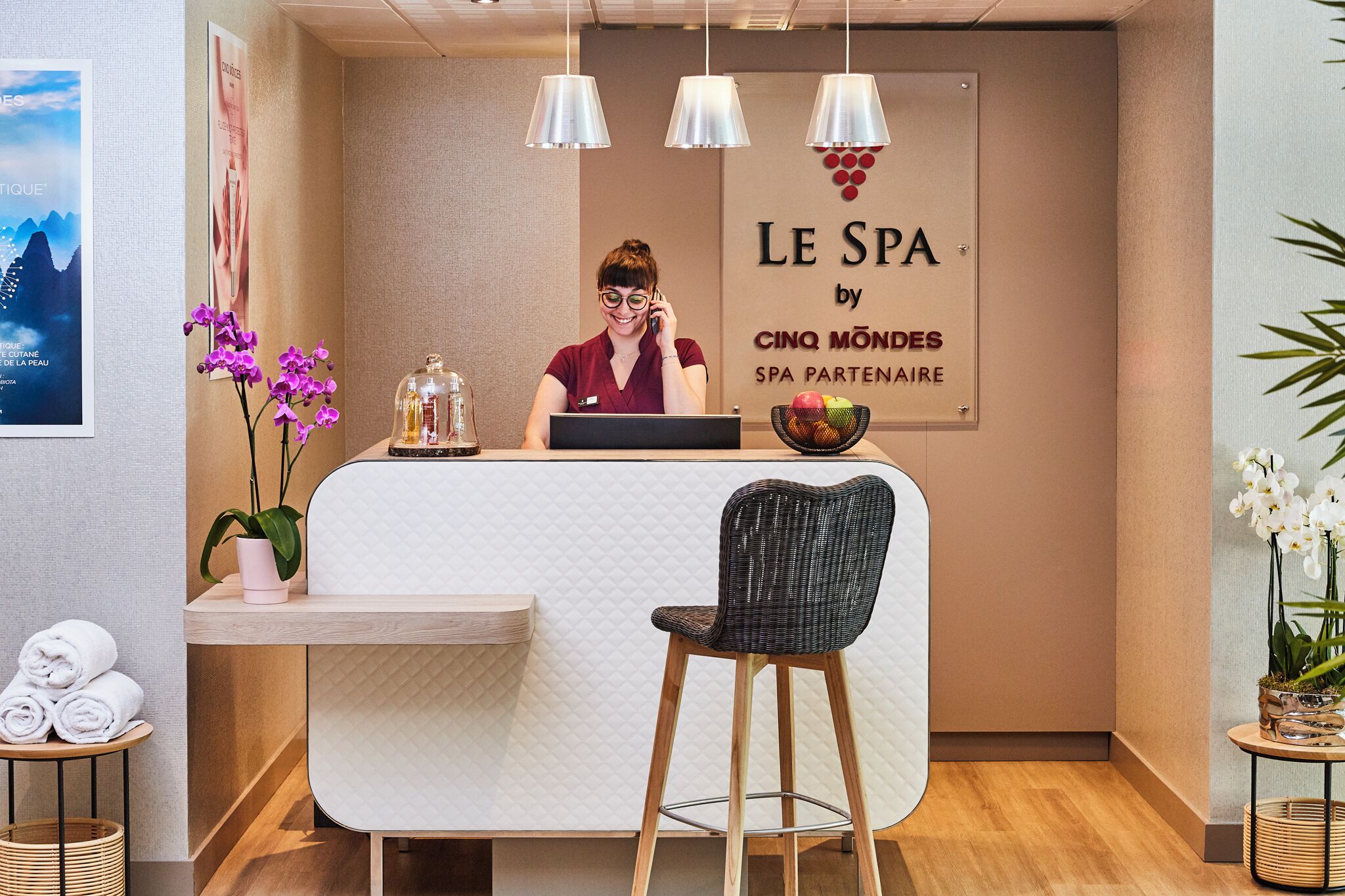 A reception area with a receptionist, a sign reading "LE SPA by CINQ MONDES SPA PARTNAIRE", a wooden reception desk, a bar stool, and a plant.