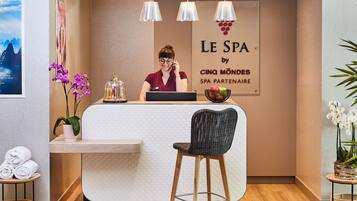 A reception area with a receptionist, a sign reading "LE SPA by CINQ MONDES SPA PARTNAIRE", a wooden reception desk, a bar stool, and a plant.