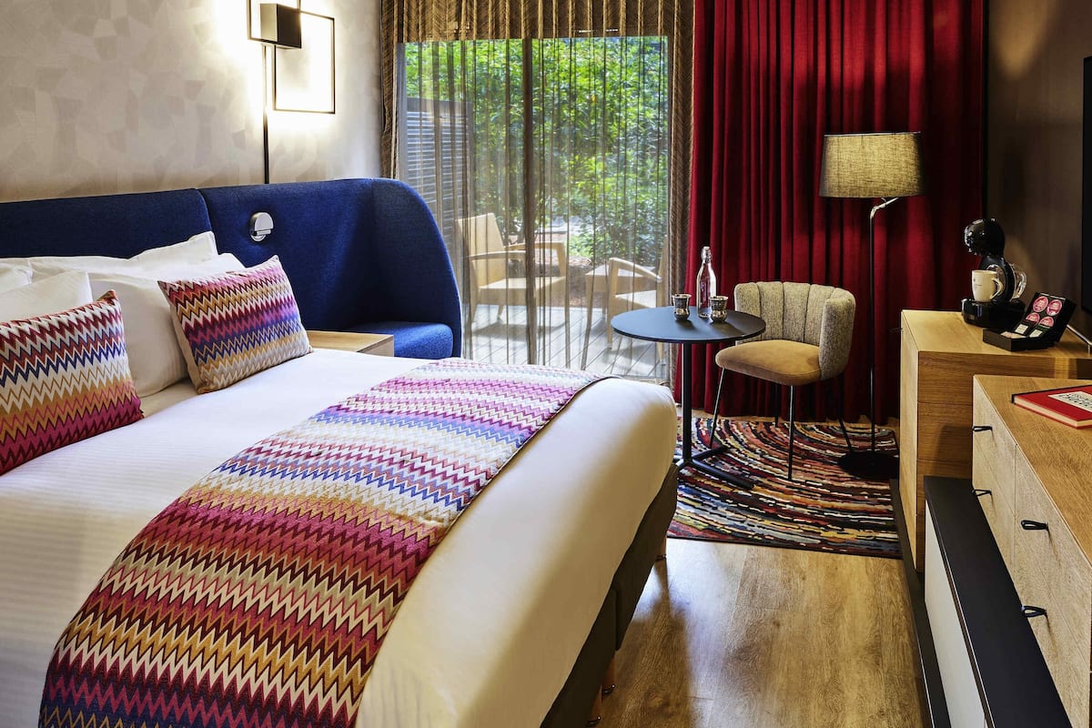 A hotel room with a large bed, a small round table, a chair, a desk with a lamp, and a view of greenery through the window.