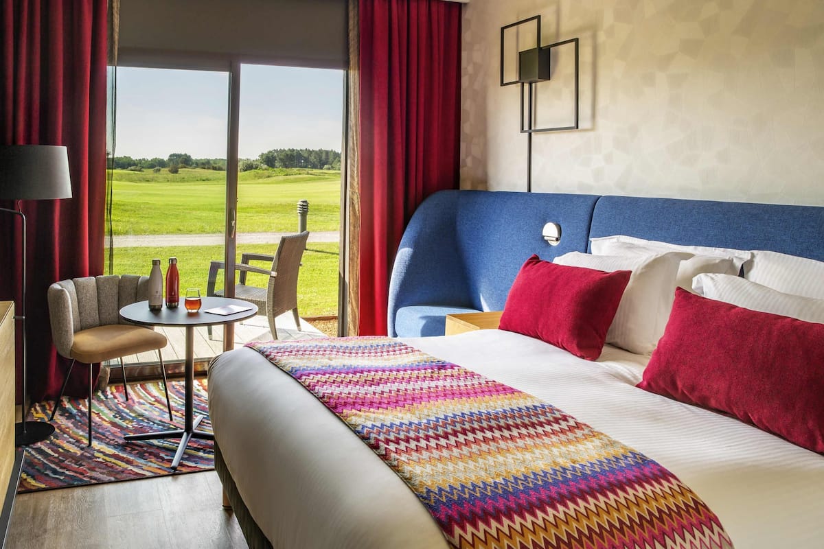 A hotel room with a large bed, a view of a golf course, a small table with a chair, and a lamp.