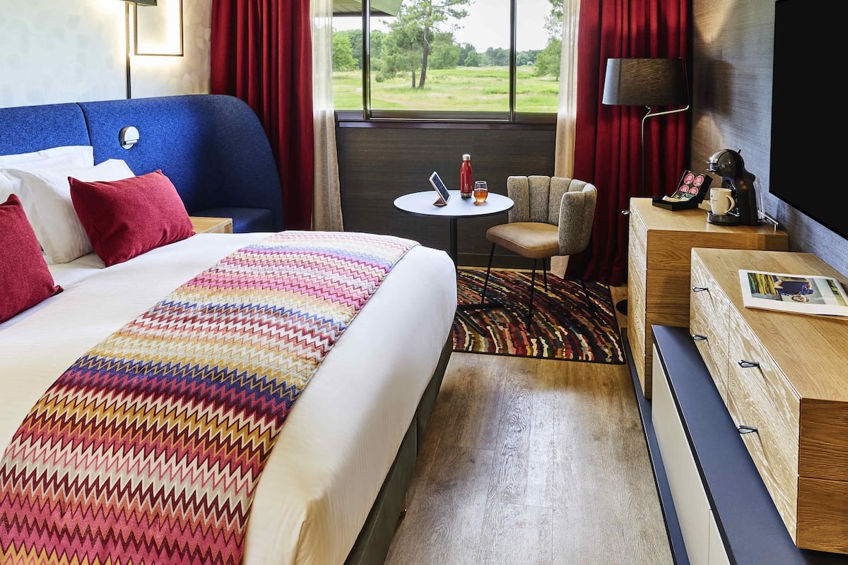 A hotel room with a large bed, a small table, a chair, a TV, and a view of the outdoors.