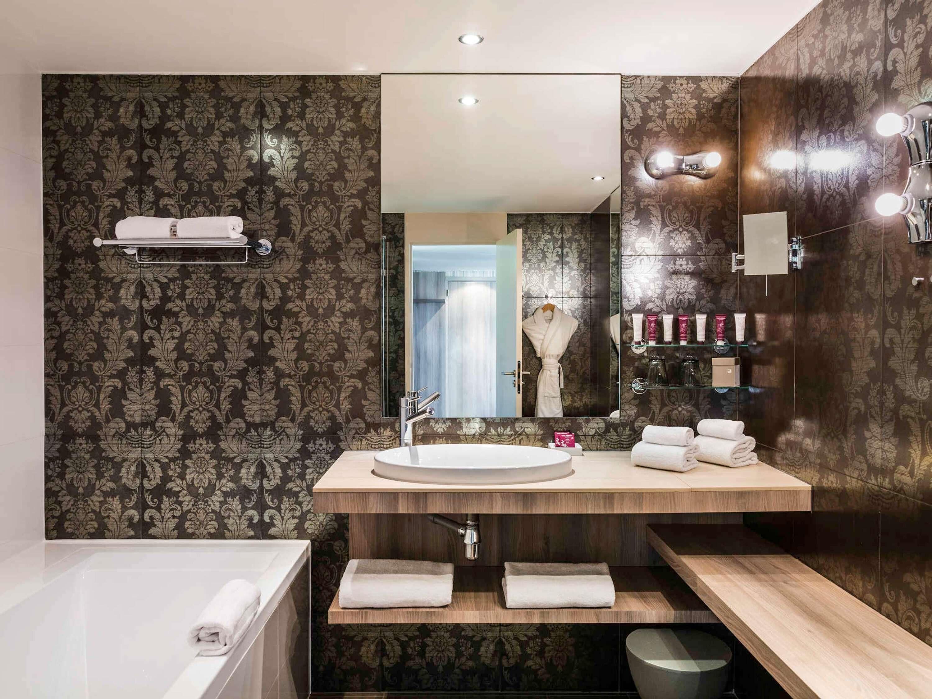 A modern bathroom with a large mirror, a round sink, and a bathtub. There are towels on the counter and a robe hanging on a rack.