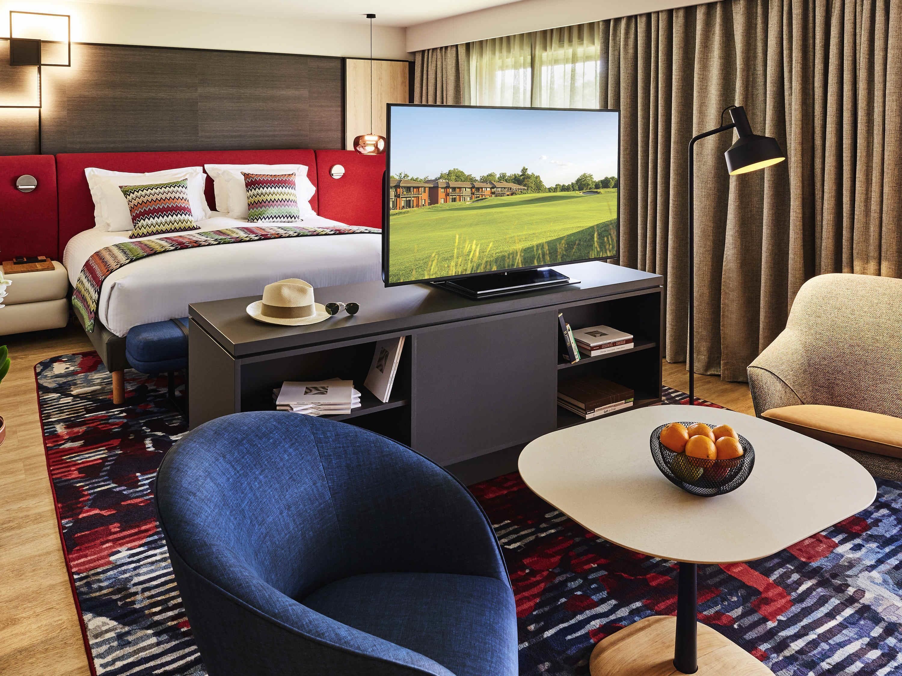 Une chambre d’hôtel comprenant un lit, une télévision, un bureau avec un ordinateur portable, une chaise, une petite table avec un panier de fruits et une bibliothèque.