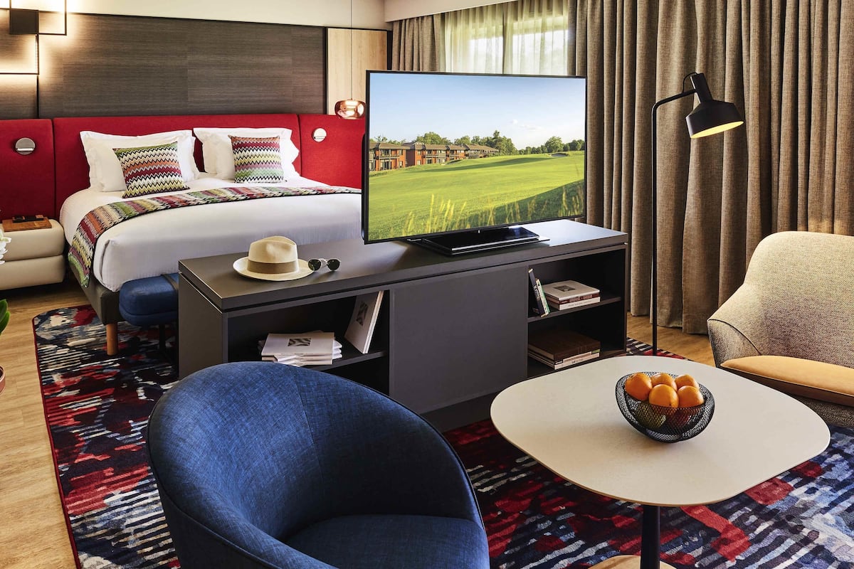 A hotel room with a bed, a TV, a desk with a laptop, a chair, a small table with a fruit bowl, and a bookshelf.