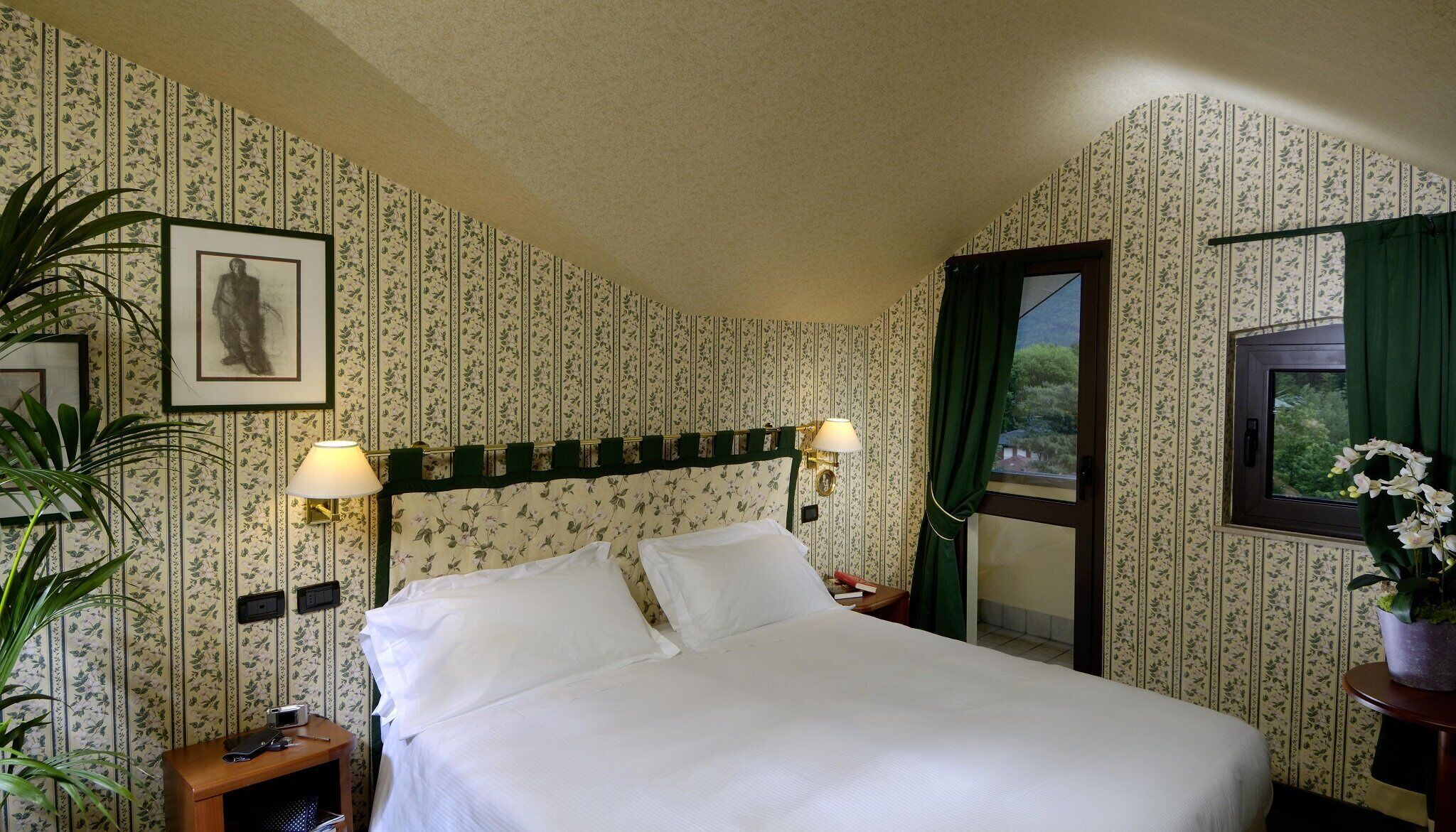 Ein ordentlich gemachtes Bett in einem Zimmer mit floralem Wandpapier, ein Nachttisch mit Lampe, ein gerahmtes Bild an der Wand und ein Fenster mit Blick auf Grünflächen.