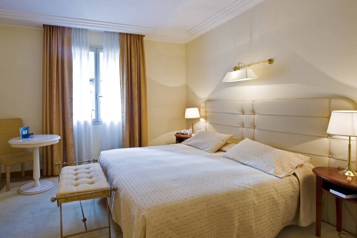 A hotel room with a bed, a small table, a chair, a TV, and a window with curtains.