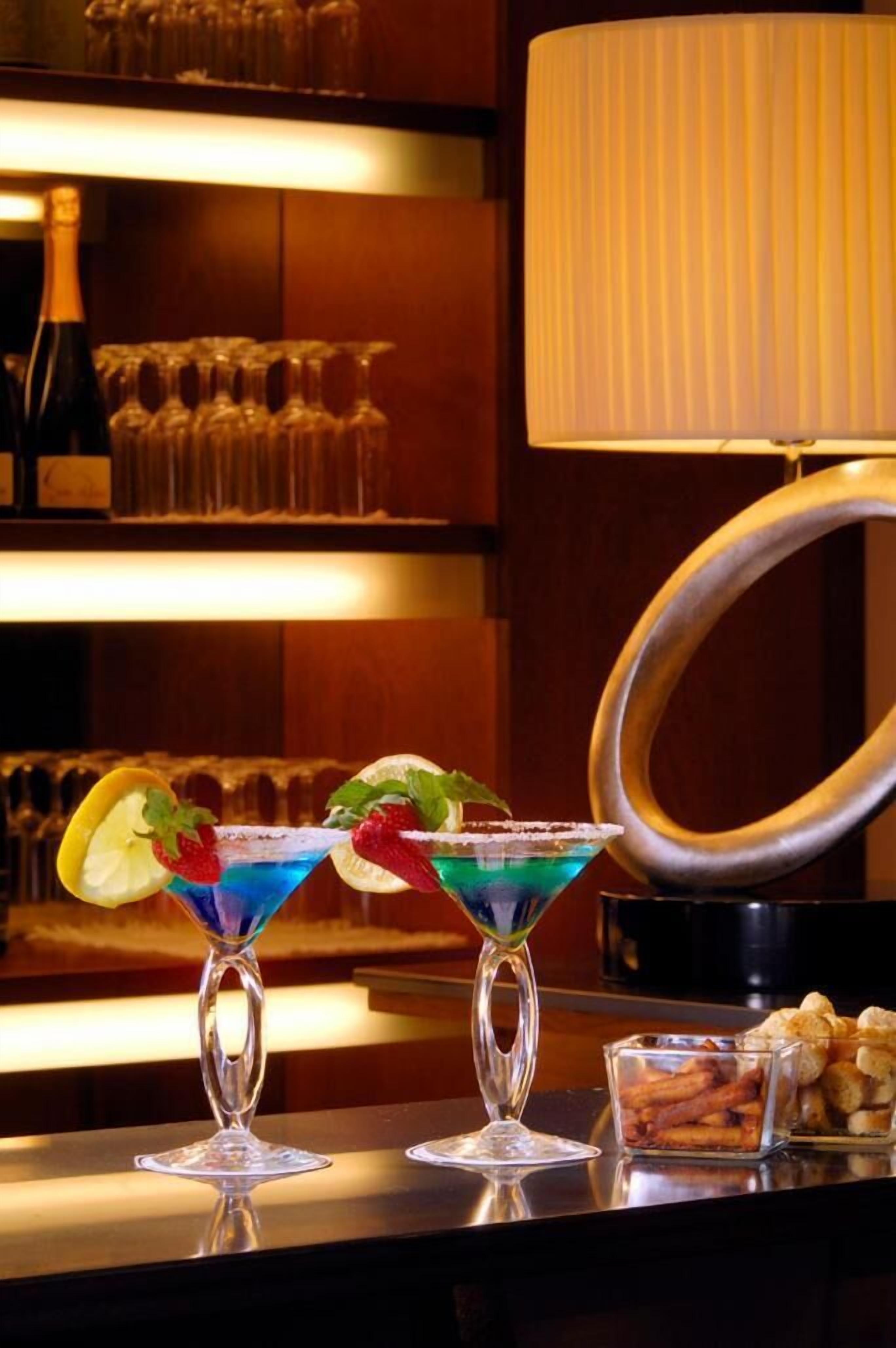 Two cocktails on a bar counter, a bottle of wine, and a lamp in the background.