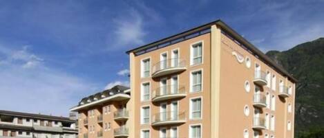A multi-story hotel with balconies and a flat roof, surrounded by greenery and mountains.
