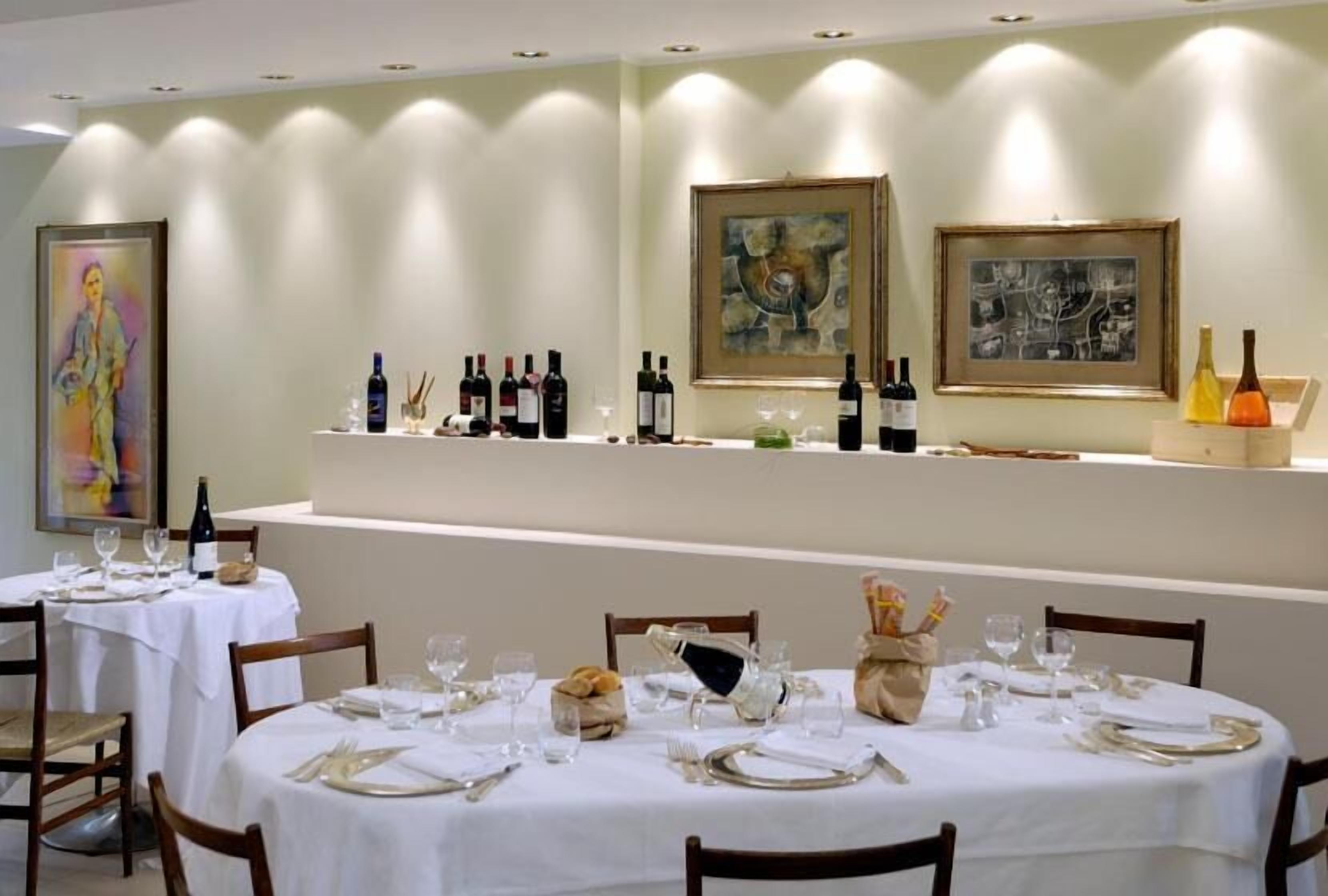 A dining area with round tables set for a meal, featuring white tablecloths, neatly arranged cutlery, and wine bottles on a shelf.